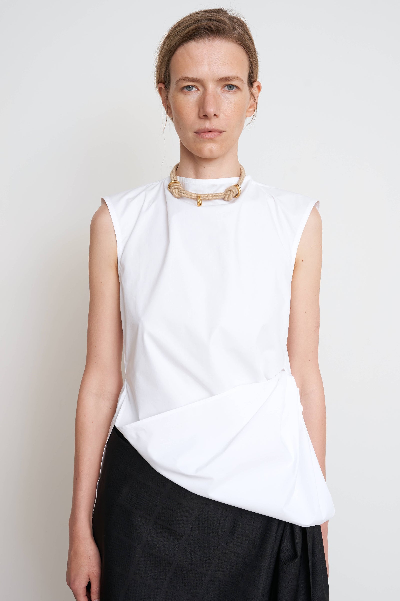 A woman wearing a white sleeveless cotton shirt with a twisted design at the waist, paired with a black skirt and a necklace. WAIST TWISTED COTTON SHIRT 